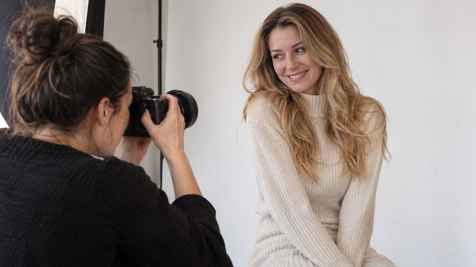 Eine Fotografin fotografiert eine blonde Frau im Strickkleid vor einem weißen Hintergrund.