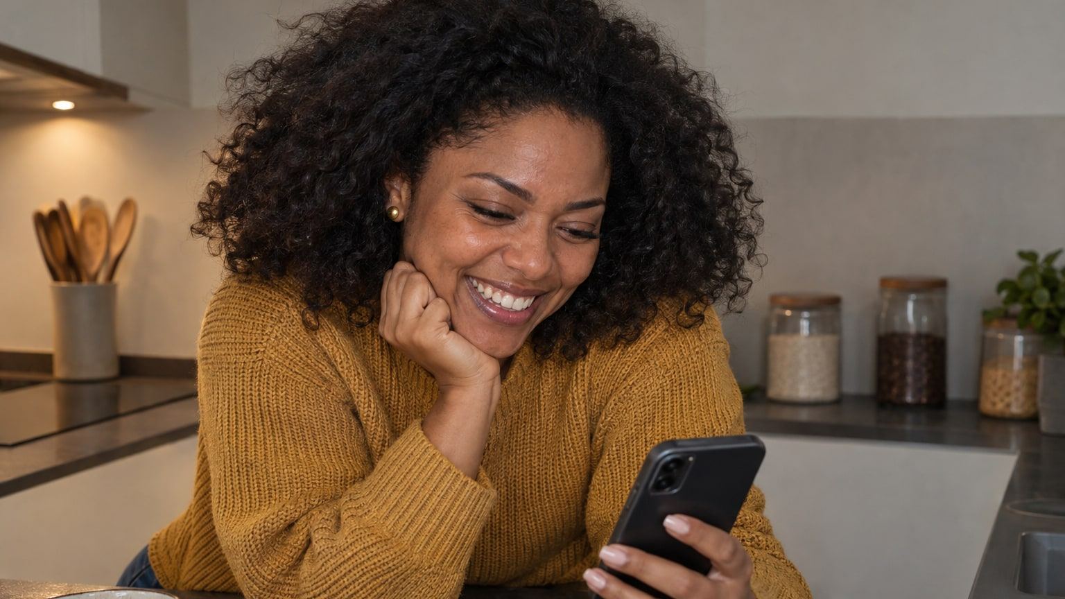 Frau mit Locken im senfgelben Pullover lacht in der Küche über ihr Handy.