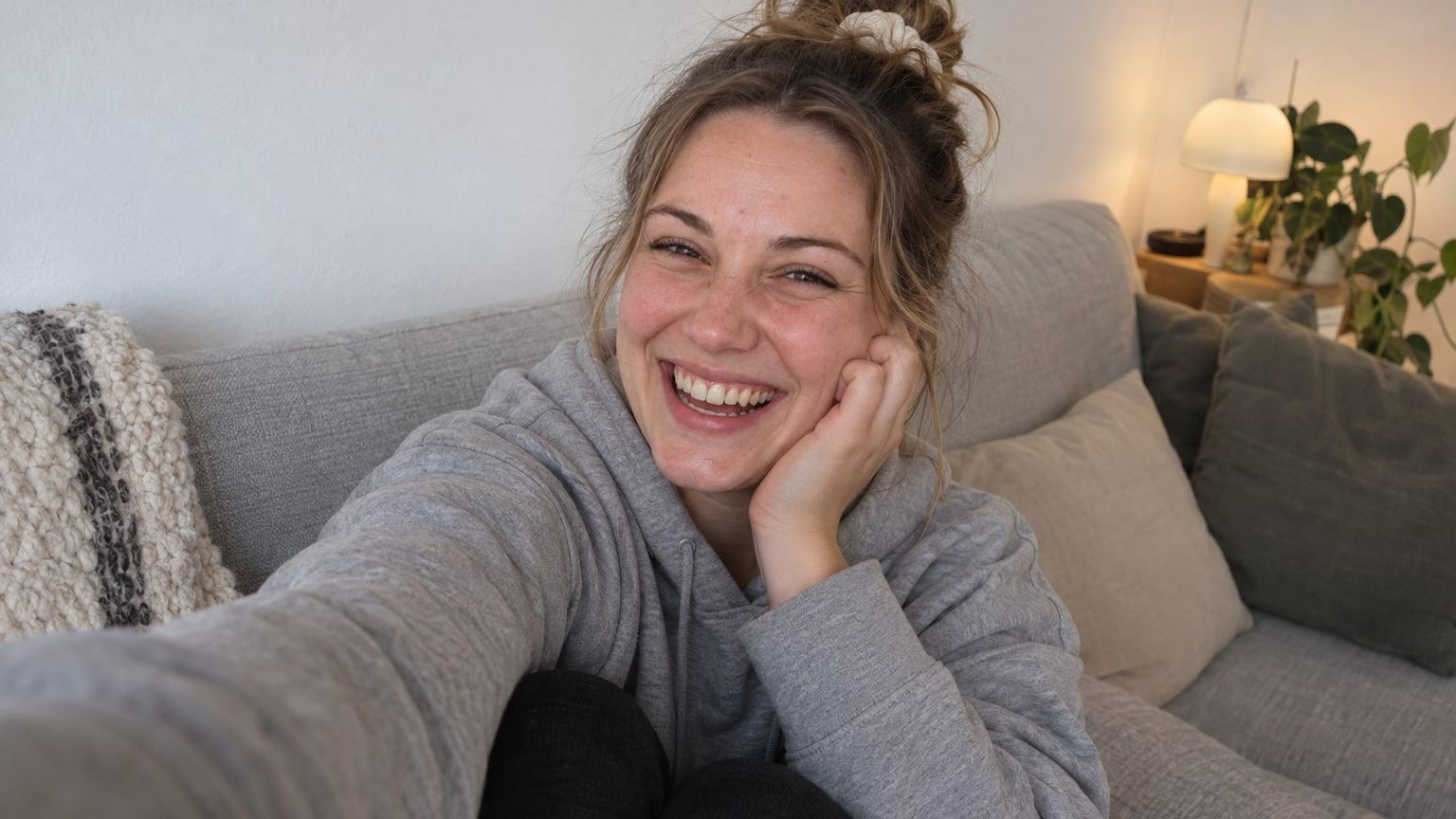 Lachende Frau im grauen Hoodie macht auf einem hellgrauen Sofa ein Selfie.
