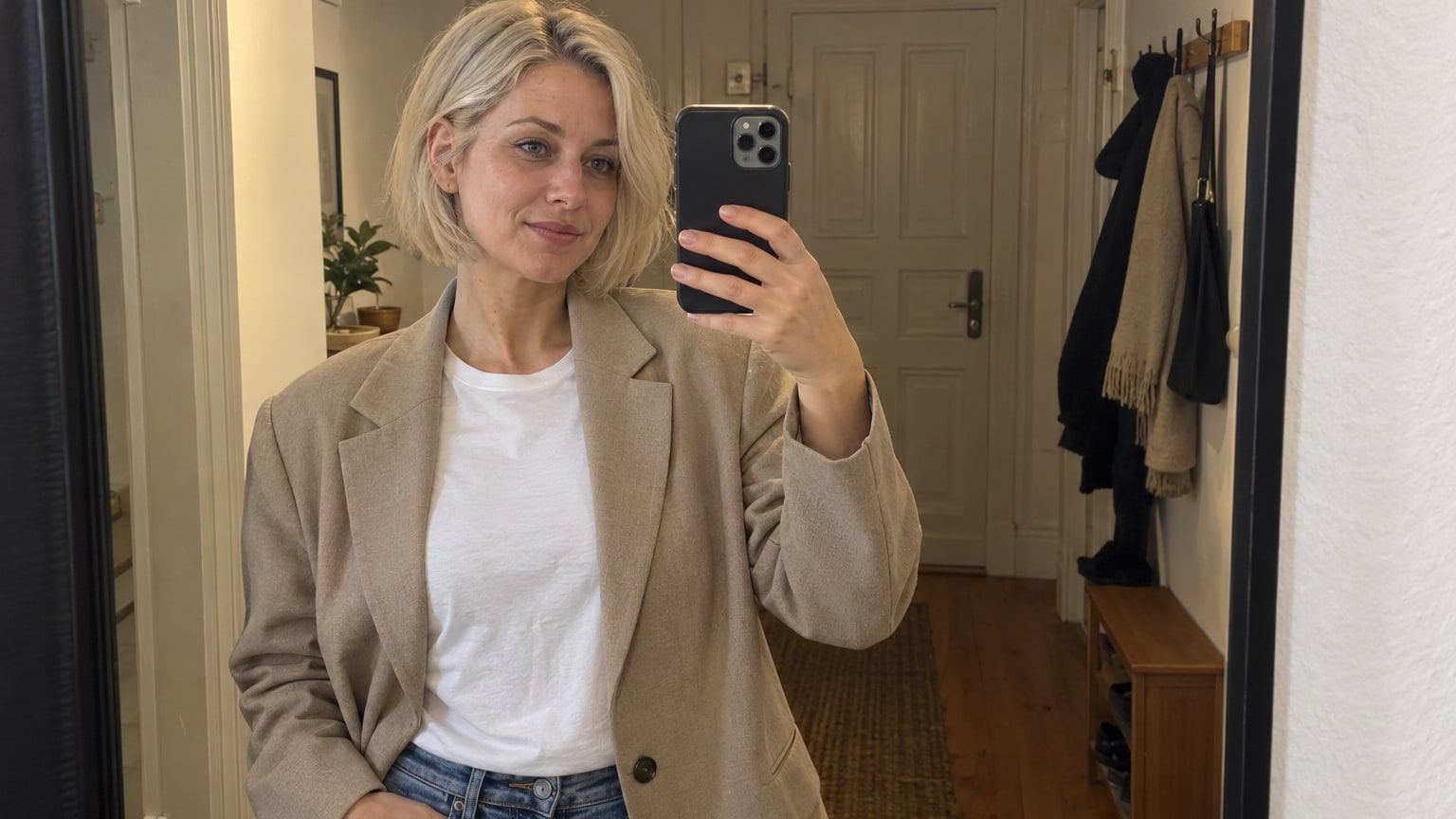 Frau mit platinblondem Bob macht im Altbauflur ein Spiegelselfie im beigen Blazer.